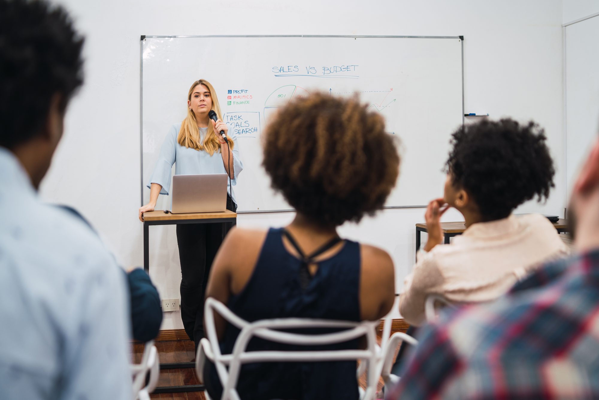 A woman at a whiteboard taking questions from colleagues seated in a meeting room
