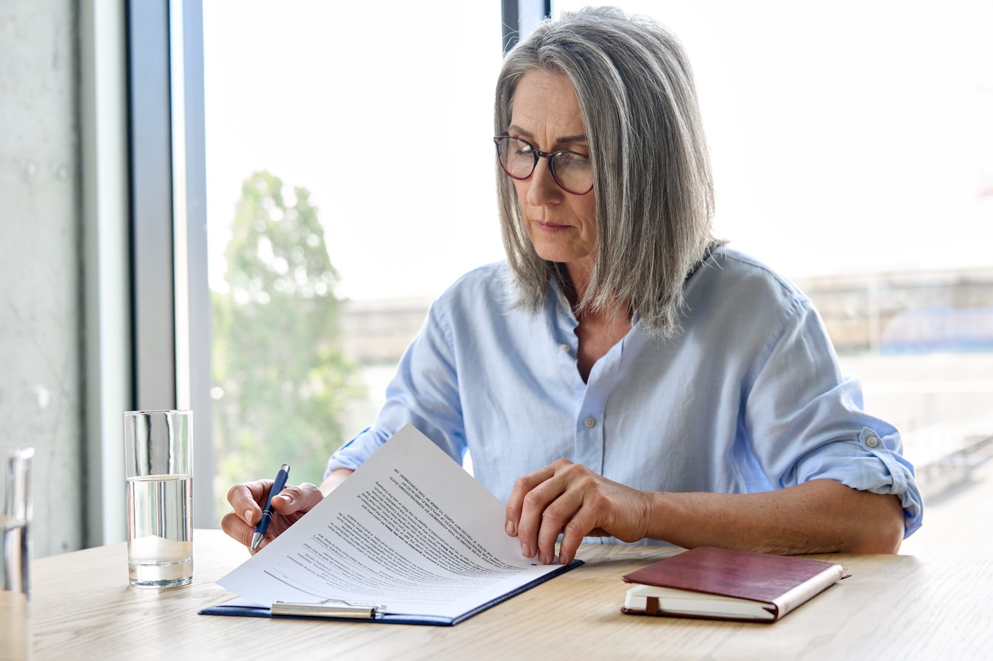 A grey-haired woman in glasses reading a contract at a desk by a window