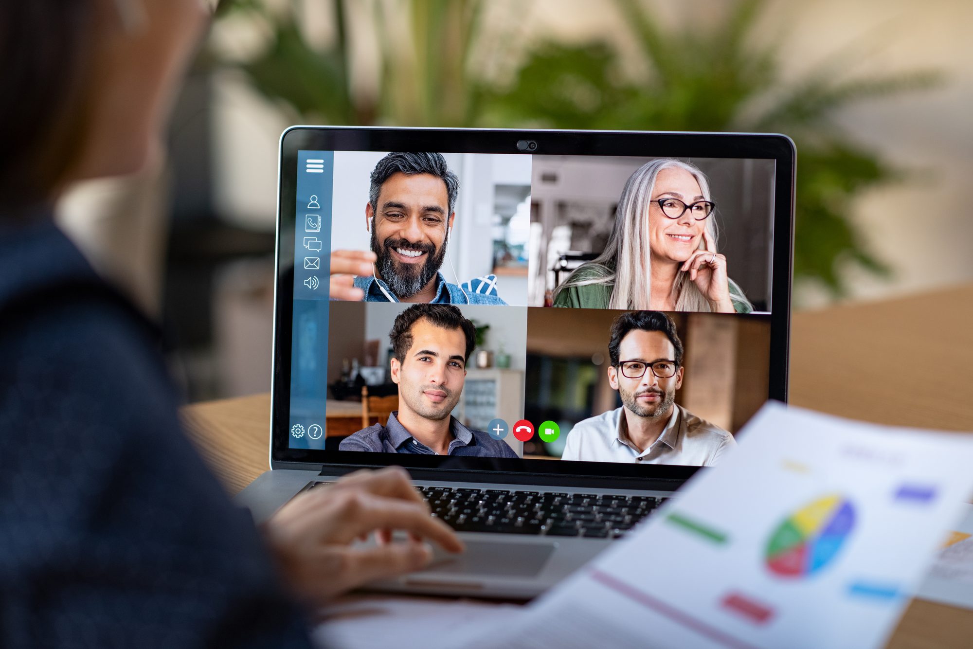 Over the shoulder of a woman on a video call with four colleagues, printed charts beside the laptop